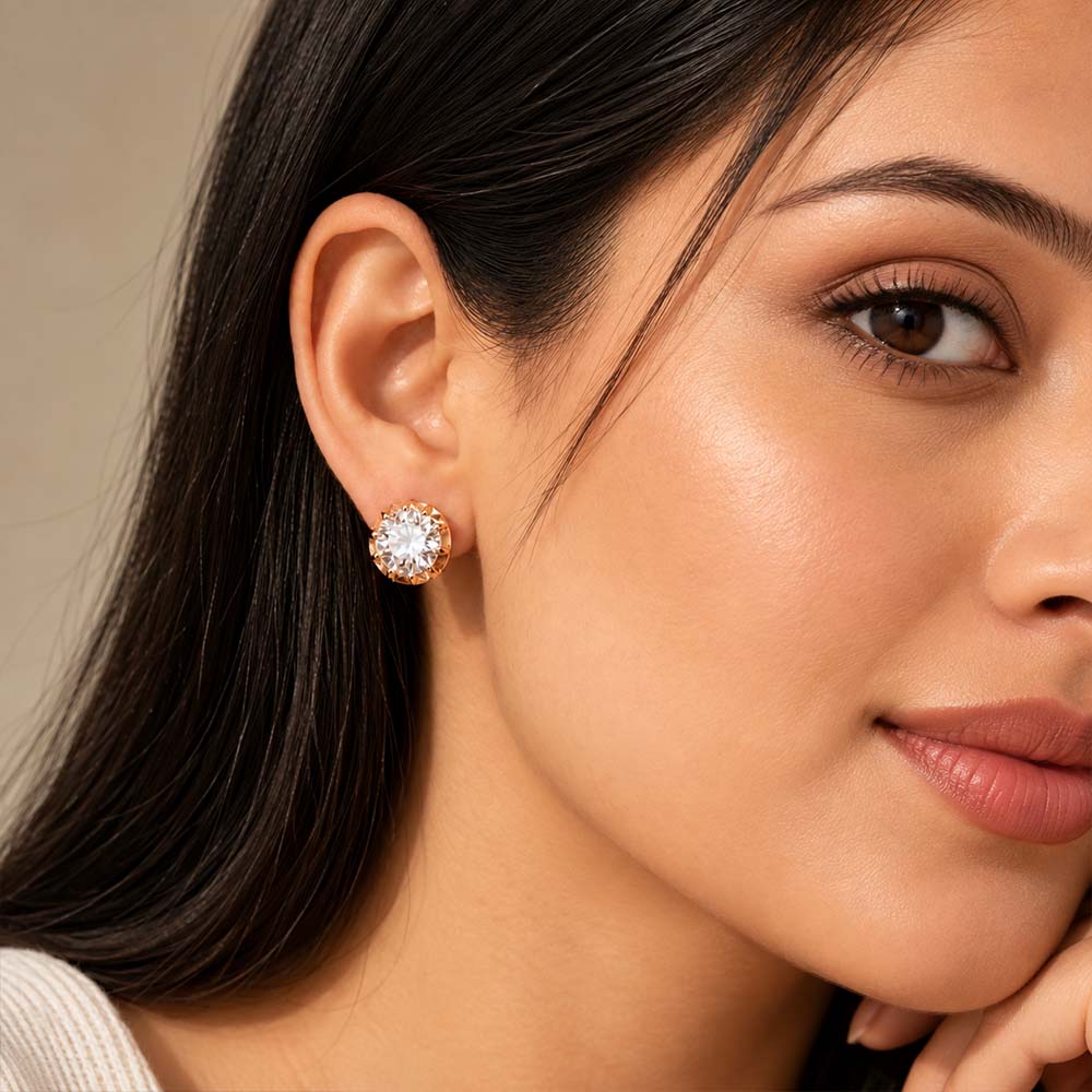 A close-up of a woman's ear wearing a single round stud earring with a clear central gemstone and a halo of small clear stones in a warm metallic setting.