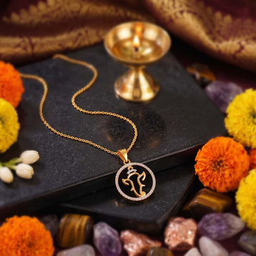 A gold necklace with a circular pendant featuring a stylized script or symbol, displayed on a black stone slab with a lit diya, flowers, and colorful stones in the background.