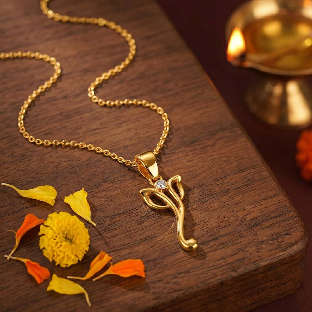Gold necklace with a leaf-shaped pendant on a wooden surface with flowers and a lamp.