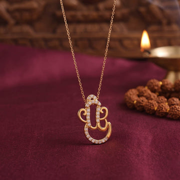 Gold Om pendant necklace adorned with small clear stones, displayed on a deep burgundy fabric surface with a blurred diya and wooden beads in the background.