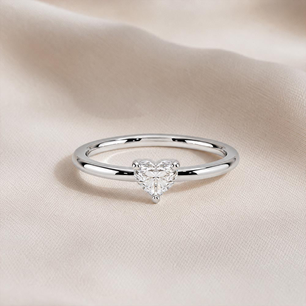 A silver ring with a heart-shaped clear gemstone, resting on a soft beige fabric background.