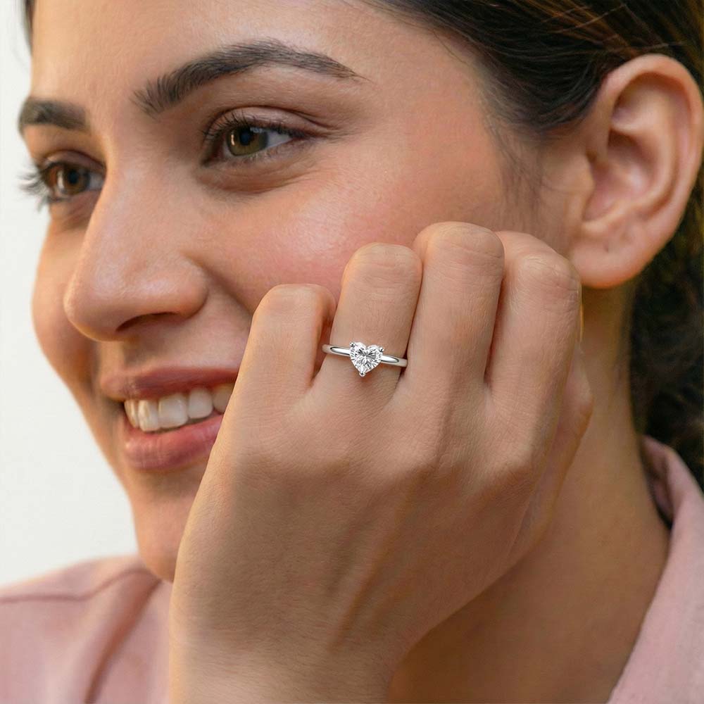 A close-up lifestyle shot of a woman wearing a delicate silver heart-shaped ring on her finger, with her hand resting near her face against a soft, light background.
