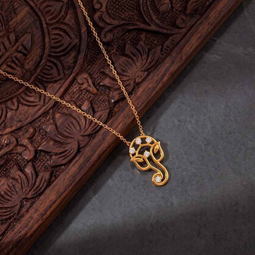 A gold-toned necklace with a Ganesha pendant, accented with small clear stones, displayed on a dark carved wooden tray over a gray surface.