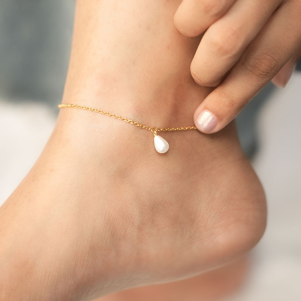 Gold anklet with a pearl charm on a person's ankle against a neutral background