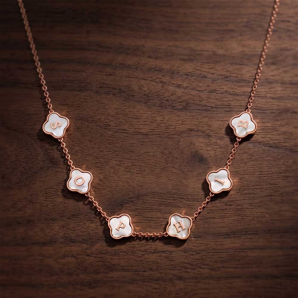 A rose gold necklace with five white enamel flower-shaped charms, each engraved with a different letter (E, Q, P, H), displayed on a dark wood background.